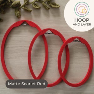 May include: Three matte scarlet red oval embroidery hoops of different sizes. Each hoop has a metal clasp and is made of a smooth material. The text "HOOP AND LAYER" is in a circle in the upper right corner. The hoops are in a wooden surface.
