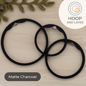 May include: Three matte charcoal embroidery hoops of different sizes are shown. The hoops are black with a metal clasp. The text "HOOP AND LAYER" is in a circle in the upper right corner. The text "Matte Charcoal" is in a dark grey rectangle at the bottom.