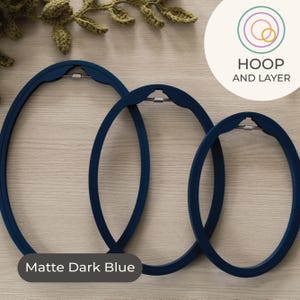 May include: Three matte dark blue oval embroidery hoops of different sizes are shown on a light wood surface. Each hoop has a metal clasp. The text "HOOP AND LAYER" is in a circle. The text "Matte Dark Blue" is at the bottom of the image.