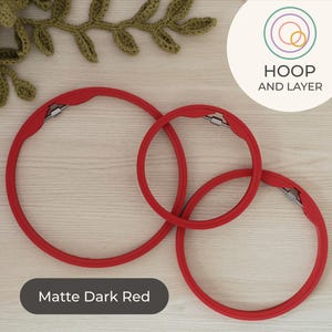 May include: Three matte dark red embroidery hoops of varying sizes are intertwined, with silver clasps. The hoops are arranged on a light wood surface, with a green crocheted leaf design in the background. The text "HOOP AND LAYER" is in a circle.