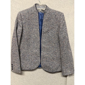 Op de afbeelding: Een grijze tweed blazer met een blauwe voering. De jas heeft een kraagloos ontwerp en drie knopen op elke manchet. De stof heeft vlekken van blauw, rood en bruin.