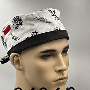 May include: A white scrub cap featuring a repeating pattern of San Antonio Spurs basketball logos and text. The cap has a solid black band along the bottom edge and a small red rectangular tag on the side. The design includes basketball graphics.