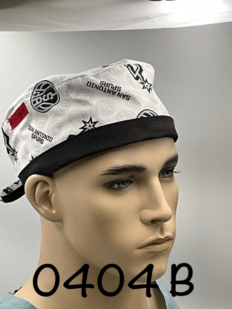May include: A white scrub cap featuring a repeating pattern of San Antonio Spurs basketball logos and text. The cap has a solid black band along the bottom edge and a small red rectangular tag on the side. The design includes basketball graphics.