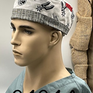 May include: A white scrub cap featuring a repeating pattern of the San Antonio Spurs basketball logo and text, accented by a grey and white textured fabric band along the bottom edge. The cap is displayed on a mannequin head wearing a light blue scrub top.