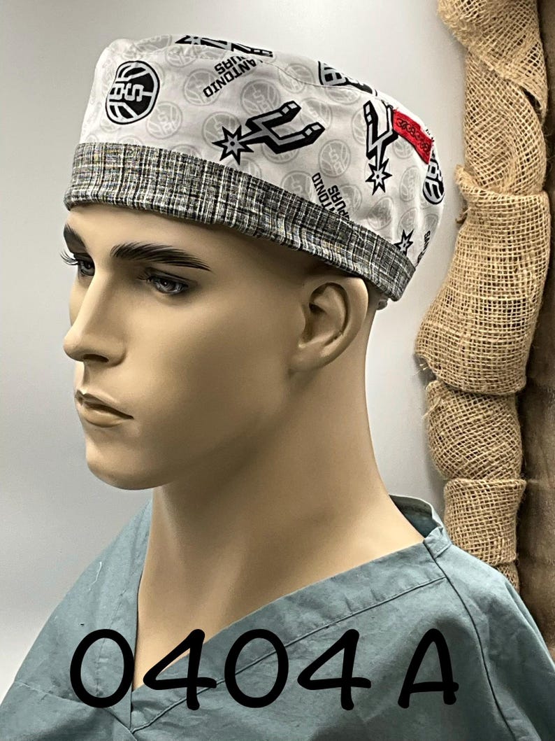 May include: A white scrub cap featuring a repeating pattern of the San Antonio Spurs basketball logo and text, accented by a grey and white textured fabric band along the bottom edge. The cap is displayed on a mannequin head wearing a light blue scrub top.