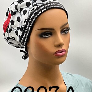 May include: A scrub cap with a repeating pattern of black and white footballs. The cap has a black band with "FIFA WORLD CUP" text. The number "0297 A" is printed on the front. The cap is displayed on a mannequin.