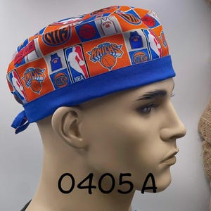 May include: A scrub cap featuring an orange background with a repeating pattern of New York Knicks basketball logos, basketballs, and jerseys. The cap has a solid blue fabric band along the bottom edge and ties at the back for an adjustable fit.