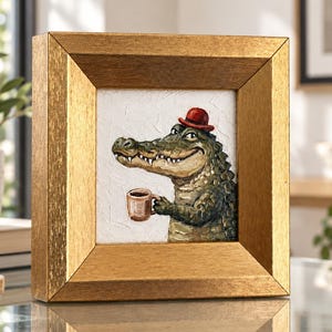 May include: A framed painting featuring a green alligator wearing a small red hat and holding a brown coffee mug. The artwork is set within a thick, textured gold frame with a square profile, showcasing a white background behind the alligator.