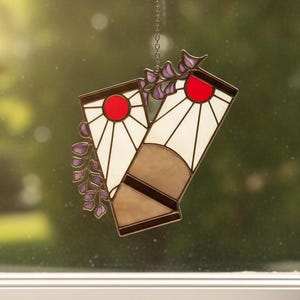 May include: A stained glass suncatcher featuring two rectangular panels with white and tan segments, each topped with a red sunburst design. The piece is accented with purple floral vines and hangs from a thin metal chain against a window.
