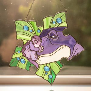 Pode incluir: Um apanhador de sol de vitral em forma de peça de puzzle com um dinossauro roxo e uma criatura mais pequena aconchegados um ao outro. O design é apresentado sobre um fundo de folhas verdes com detalhes circulares azuis e uma corrente.