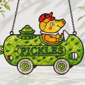 Op de afbeelding: Een glas-in-lood suncatcher in de vorm van een groene augurk-auto met een geel varken met een rode pet aan het stuur. Het woord PICKLES staat in het midden van het groene lichaam. Het object heeft een zwart metalen frame en twee zwarte kettingen.