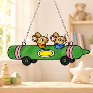May include: A stained glass suncatcher featuring two brown mice with pink ears riding in a green crayon-shaped car. The car has black wheels, pink accents on the ends, and a yellow oval in the centre. It hangs from a silver metal chain.