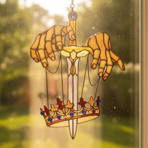 May include: A stained glass suncatcher featuring two pale yellow skeletal hands holding thin black strings attached to a crown. A central sword with a white blade and a blue gem at the hilt sits between the hands, pointing down through the crown.
