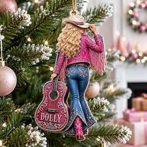 Könnte beinhalten: Ein Weihnachtsbaumschmuck, der eine blonde Figur mit Cowboyhut, einer leuchtend pinken Fransenjacke und einer blauen Jeans zeigt. Die Figur hält eine pinke Akustikgitarre, auf deren Korpus der Name DOLLY in weißen Buchstaben aufgedruckt ist.