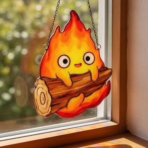 Könnte beinhalten: Ein Buntglas-Sonnenfänger in Form einer Cartoon-Flammenfigur mit großen weißen Augen und einem kleinen Lächeln, die einen braunen Holzscheit hält. Die Flamme ist in leuchtenden Gelb-, Orange- und Rottönen gehalten und an einer dünnen Metallkette aufgehängt.