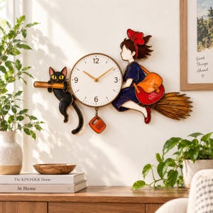 Puede incluir: Un reloj de pared con esfera circular blanca y manecillas doradas, flanqueado por un gato negro a la izquierda y una figura con lazo rojo y bolso naranja montada en una escoba a la derecha, con un pequeño colgante naranja en la base.