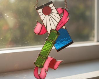 Puede incluir: Un atrapasueños de vitral que presenta un panel rectangular blanco con un diseño de sol rojo, un segmento verde con textura de bambú, una sección rectangular azul y acentos rosados en forma de cinta, unidos por líneas de soldadura de cobre.