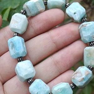 May include: A bracelet featuring light blue, faceted gemstone beads. The beads are strung together with small, dark metal spacers. The bracelet is held in a hand, showcasing the color and texture of the stones.