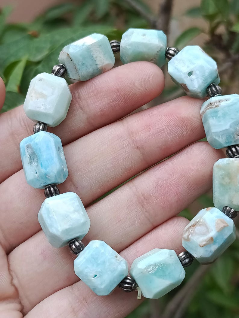 May include: A bracelet featuring light blue, faceted gemstone beads. The beads are strung together with small, dark metal spacers. The bracelet is held in a hand, showcasing the color and texture of the stones.