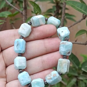 May include: A light blue gemstone bracelet with faceted, rectangular beads. The beads are strung together with dark metal connectors. The bracelet is held against a background of green foliage.