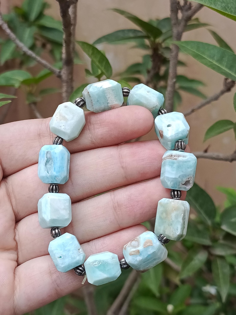 May include: A light blue gemstone bracelet with faceted, rectangular beads. The beads are strung together with dark metal connectors. The bracelet is held against a background of green foliage.
