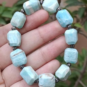 May include: A light blue gemstone bracelet. The bracelet features faceted, rectangular beads in varying shades of blue and white, strung together with small, dark metal beads. The bracelet is held in a hand, showcasing its color and design.