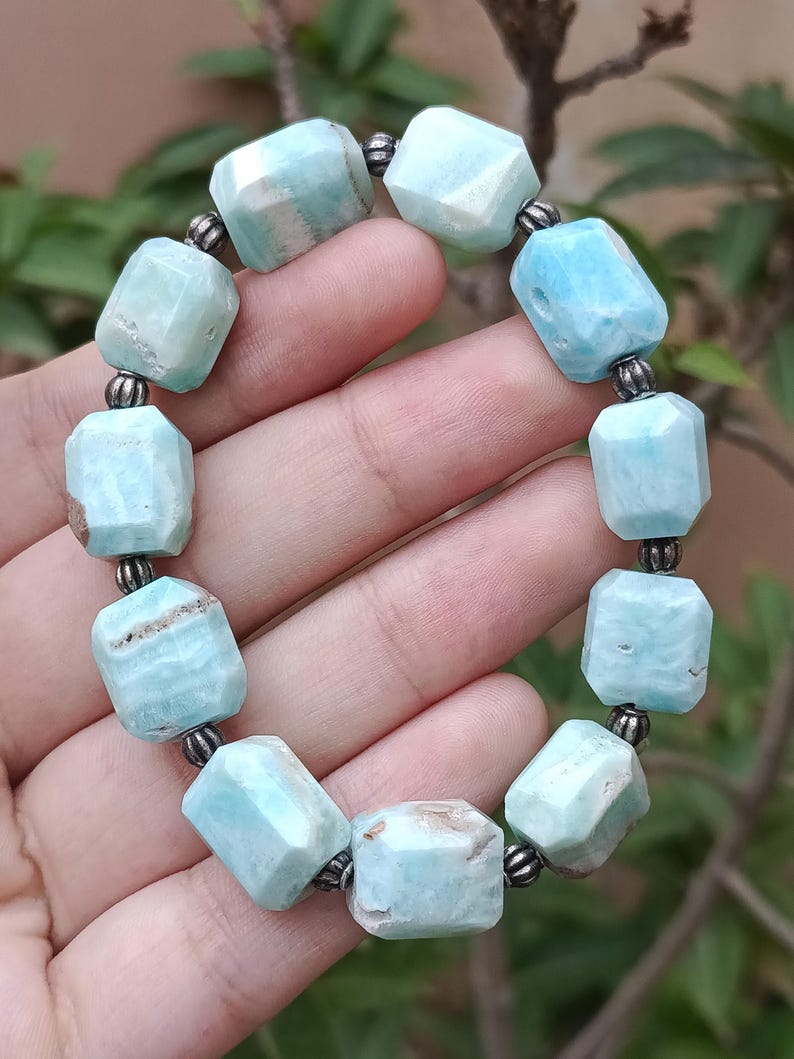 May include: A light blue gemstone bracelet. The bracelet features faceted, rectangular beads in varying shades of blue and white, strung together with small, dark metal beads. The bracelet is held in a hand, showcasing its color and design.