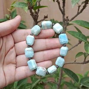 May include: A light blue gemstone bracelet with a series of rectangular beads. The beads are connected by small, dark gray metal links. The bracelet is held by a hand in front of a background of green leaves and branches.