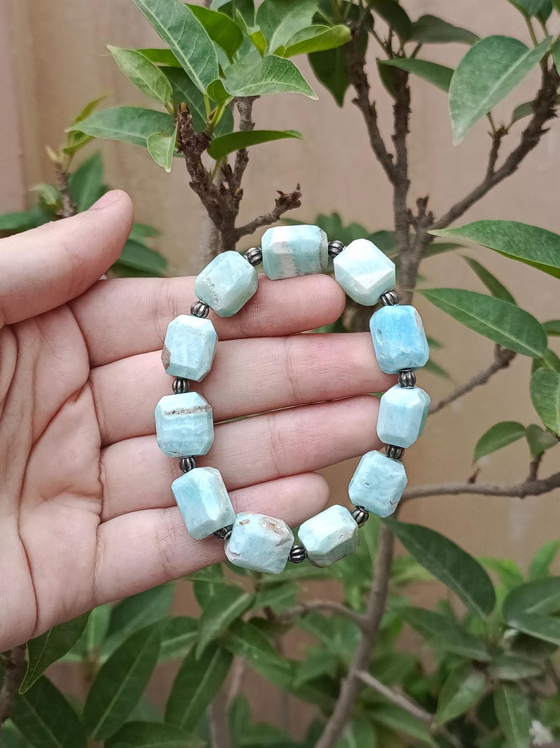 May include: A light blue gemstone bracelet with a series of rectangular beads. The beads are connected by small, dark gray metal links. The bracelet is held by a hand in front of a background of green leaves and branches.