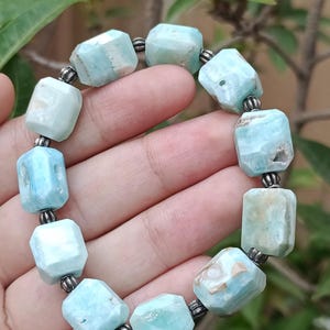 May include: A light blue gemstone bracelet with faceted rectangular beads. The beads are strung together with small silver-colored spacers. The bracelet is held in a hand, with a blurred background of green leaves and branches.
