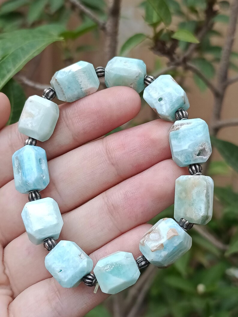 May include: A light blue gemstone bracelet with faceted rectangular beads. The beads are strung together with small silver-colored spacers. The bracelet is held in a hand, with a blurred background of green leaves and branches.