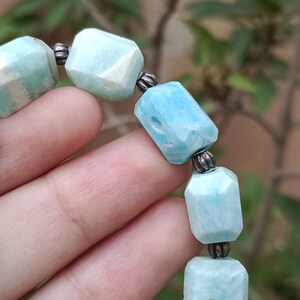 May include: A close-up of a bracelet featuring light blue, faceted gemstone beads. The beads are connected by small, silver-colored metal beads. The bracelet is displayed against a blurred green background.