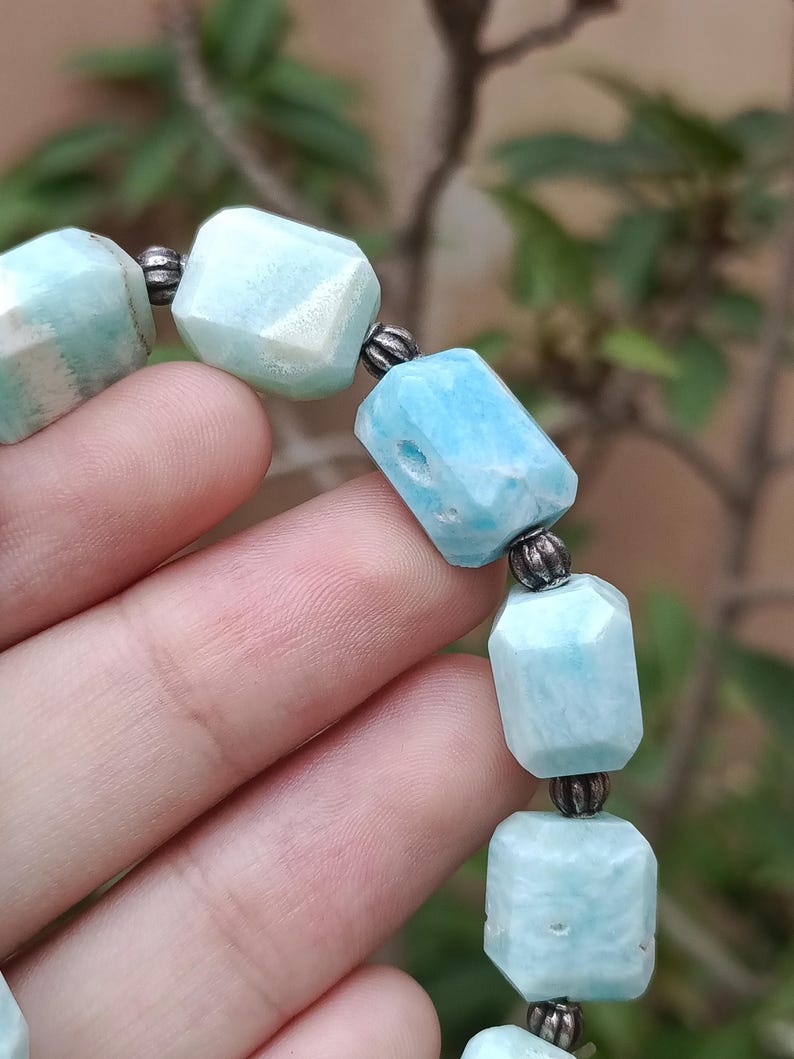 May include: A close-up of a bracelet featuring light blue, faceted gemstone beads. The beads are connected by small, silver-colored metal beads. The bracelet is displayed against a blurred green background.