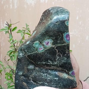 May include: A dark green stone sculpture with a polished surface. The stone features patches of teal and purple, with a natural, organic shape. The sculpture is held in a person's hand.