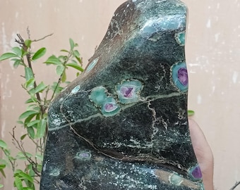 Op de afbeelding: Een donkergroene stenen sculptuur met een gepolijst oppervlak. De steen heeft vlekken van teal en paars, met een natuurlijke, organische vorm. De sculptuur wordt in de hand van een persoon gehouden.