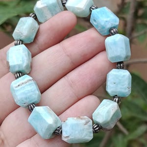 May include: A light blue gemstone bracelet. The bracelet is made of faceted, rectangular beads with a pale blue color and some brown inclusions. The beads are strung together with small, silver-colored metal beads.