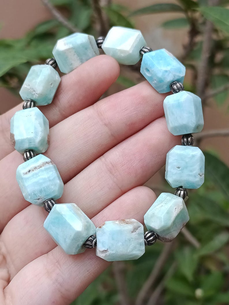 May include: A light blue gemstone bracelet. The bracelet is made of faceted, rectangular beads with a pale blue color and some brown inclusions. The beads are strung together with small, silver-colored metal beads.