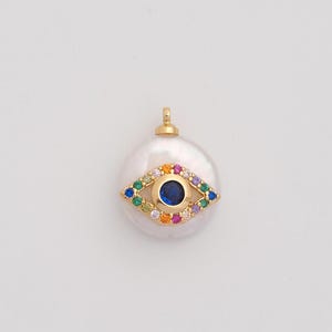 May include: A gold-colored pendant with a large, white, oval-shaped pearl. The pendant features a colorful eye design with a blue center and surrounding gemstones in various colors. The pendant has a small loop at the top for a chain.