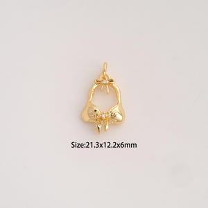 Könnte beinhalten: Ein goldfarbener Metallanhänger in Form eines Bikini-Oberteils mit einer kleinen Schleife in der Mitte und einer Schleife an der oberen Öse, besetzt mit zwei winzigen klaren Steinen. Der Anhänger misst 21,3 x 12,2 x 6 mm, siehe Text.