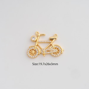 May include: A gold-colored bicycle charm with clear crystal accents. The bicycle has a round crystal on the front and crystal-studded wheels. The text "Size: 19.7x26x3mm" is below the charm.