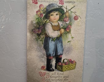 May include: Vintage Thanksgiving postcard featuring a young child in a hat and overalls holding a branch with apples. The postcard has the text "Thanksgiving Greetings" and "With all kind thoughts and Best wishes for Thanksgiving Day."