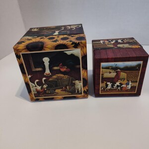 May include: Two decorative boxes with farm scenes. One box features sunflowers and a barn scene with a cow, rooster, and other farm animals. The other box is burgundy with a farm scene of cows and a woman.