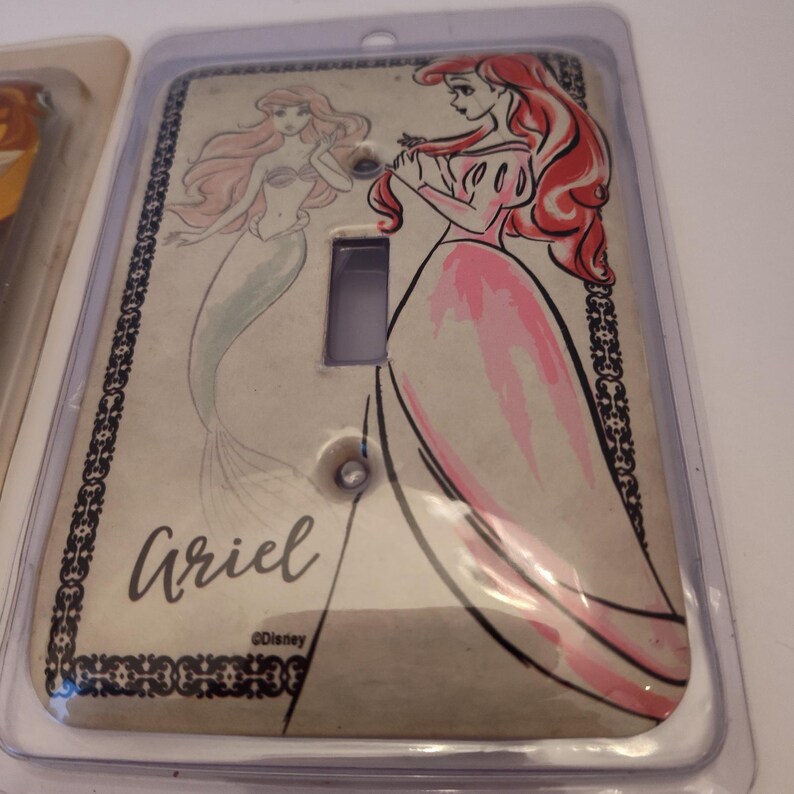 Pu&ograve; includere: Copri interruttore Ariel con un'illustrazione rosa e rossa di Ariel de La Sirenetta di Disney. La copertura ha un'estetica vintage con il nome "Ariel" in corsivo e il logo Disney.