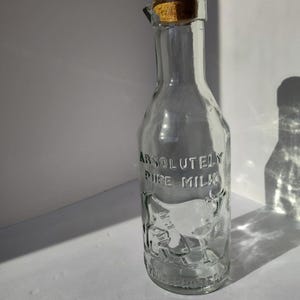 May include: A clear glass milk bottle with a cork stopper. The bottle has the words "ABSOLUTELY PURE MILK" embossed on the front, along with a cow illustration. The bottle is empty and sits on a white surface.