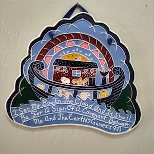 May include: A ceramic wall hanging with a blue, red, and yellow design depicting Noah's Ark. The artwork includes a rainbow, animals, and the text "I Set My Bow In The Cloud...Genesis 9:13". It has a scalloped edge and a blue ribbon for hanging.
