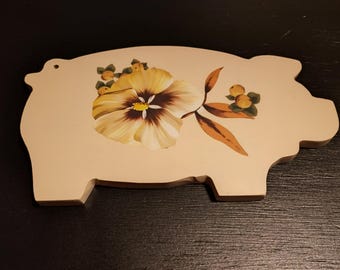 May include: A cream-colored, pig-shaped wooden cutting board. The board features a hand-painted floral design with a large yellow and white flower, brown leaves, and small orange fruits. The board has a smooth surface and is likely used for food preparation.