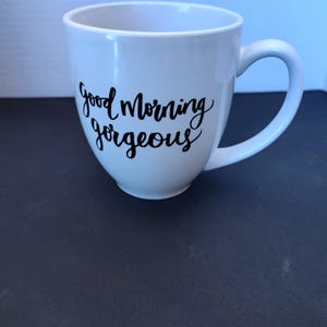 May include: White ceramic coffee mug with a curved handle. The mug features the phrase "Good Morning Gorgeous" in black script. The mug is on a dark surface.