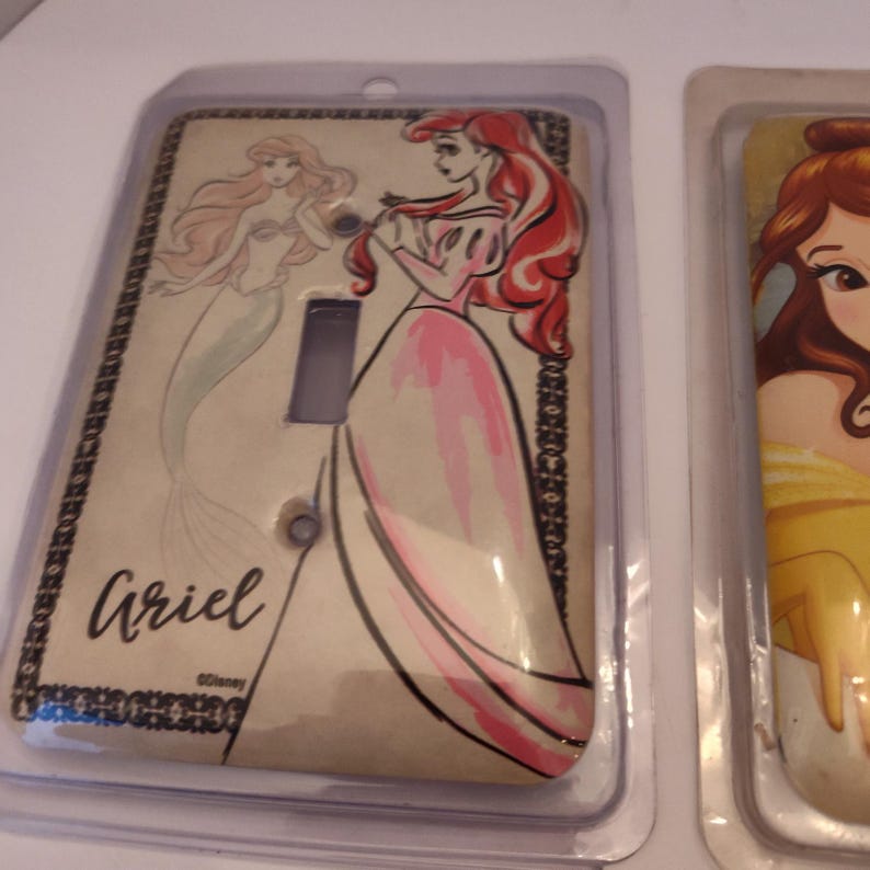 Pu&ograve; includere: Copri interruttore a tema Ariel, con un disegno di Ariel in abito rosa e come sirena. La copertura ha uno sfondo beige con bordi decorativi neri e il nome "Ariel" in corsivo.