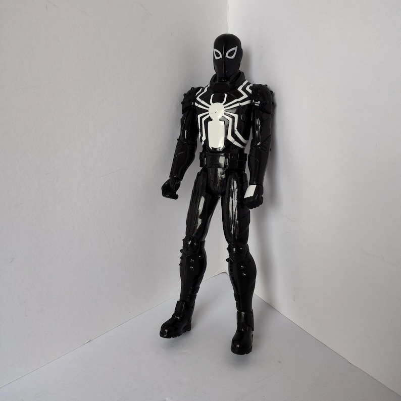 Op de afbeelding: Zwart actiefiguur van een superheld met een wit spinnenembleem op de borst. Het figuur draagt een zwart pak met witte accenten en een masker. Het figuur staat in een hoek.