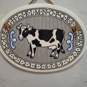 May include: Oval ceramic sign featuring a black and white cow illustration. The sign has a black and white striped background, with the alphabet and numbers around the border. Two brown wooden pieces are attached to the top.